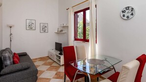 A compact living space with a glass dining table, a sofa, a TV, and a clock on the wall. - Apartments Franka - One Bedroom Apartment With Terrace and Garden View (Dubrovnik)