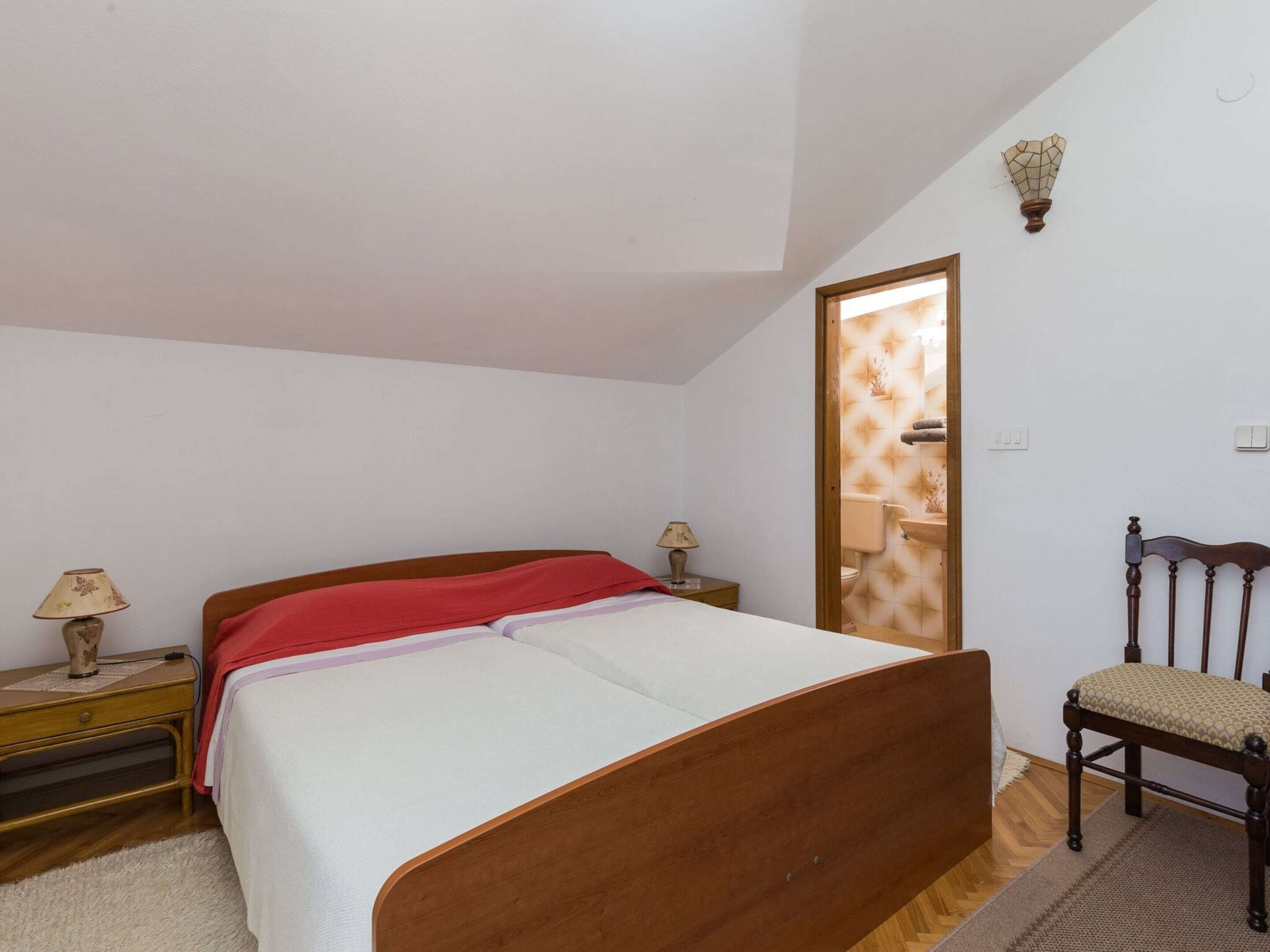 Un dormitorio con cama de madera, un mesita de noche con lámpara, una silla y un baño visible a través de una puerta.