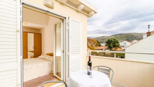 A balcony with a table and a bottle, a bedroom with a bed and a wardrobe, and a view of houses and hills in the background. - Apartments Nikola - One Bedroom Apartment With two Balconies and Sea View (Dubrovnik)
