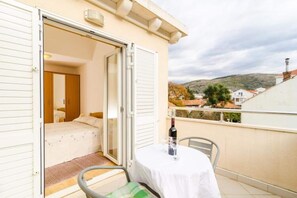 Un balcon avec une table et une bouteille, une chambre avec un lit et une armoire, et une vue sur des maisons et des collines en arrière-plan.