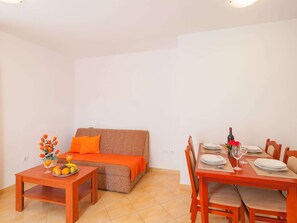 Um espaço de estar compacto com sofá, uma mesa de jantar posta para dois e uma mesinha lateral com frutas e um vaso.