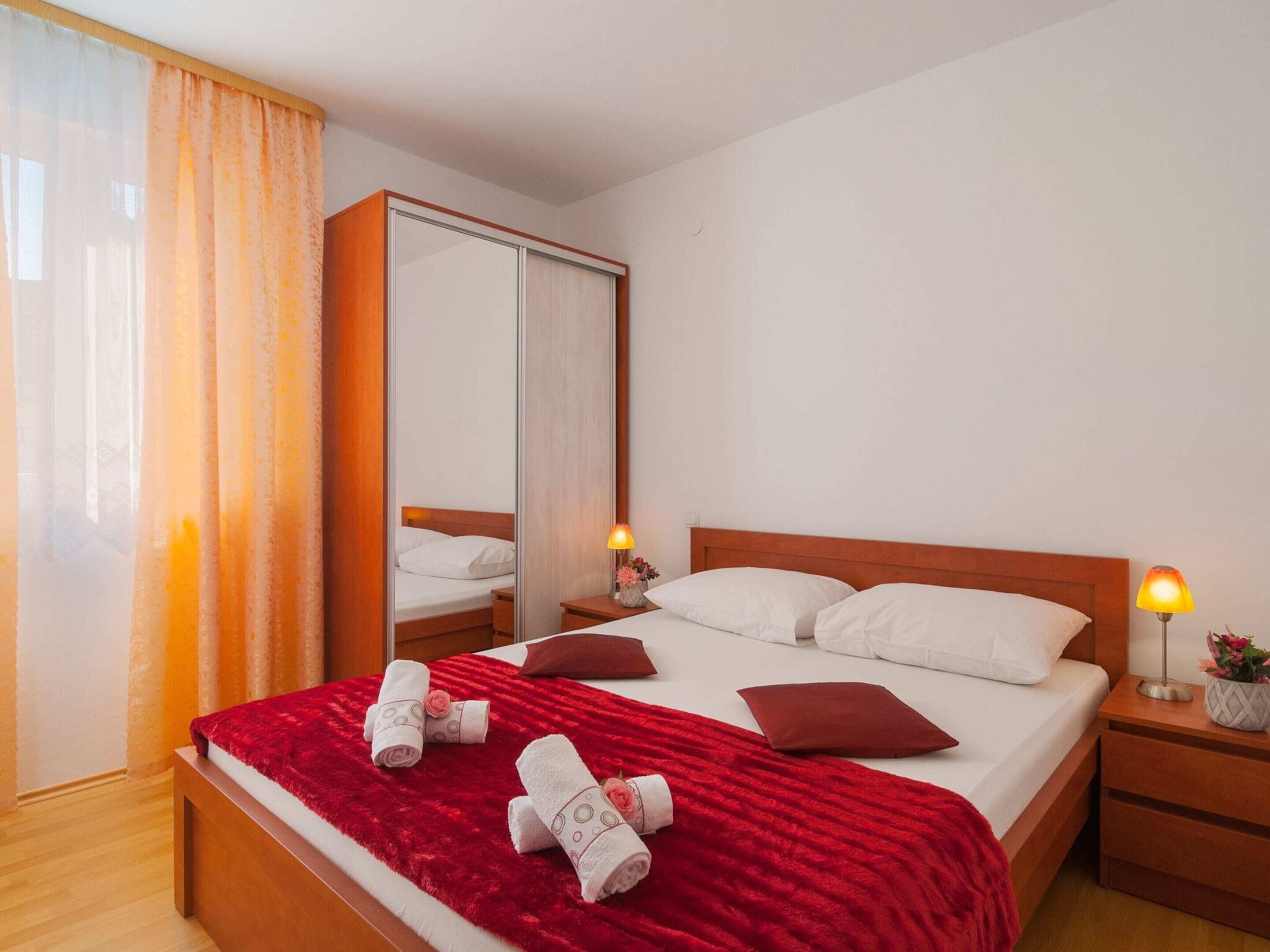 Ein Hotelzimmer mit Bett, Nachttisch, Spiegel, Fenster mit Vorhängen und einer roten Tagesdecke.
