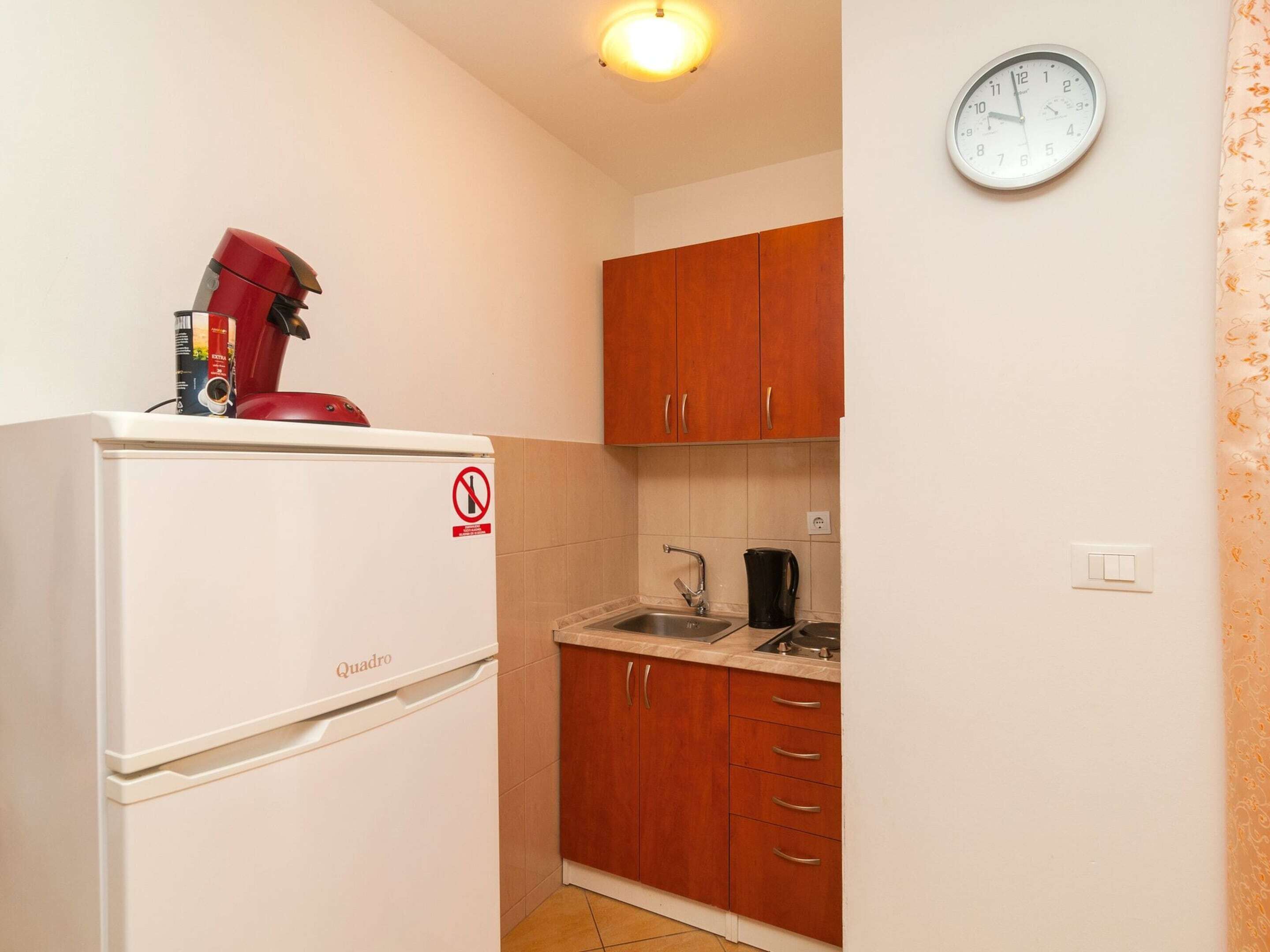 Une cuisine de taille réduite avec un réfrigérateur blanc, des placards en bois, un évier et une horloge au mur.
