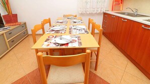 A dining area with a table set for four, wooden chairs, a kitchen with wooden cabinets, a sink, and a framed picture on the wall. - Apartments Marko Peljesac - Two Bedroom Apartment With Balcony (Orebic)