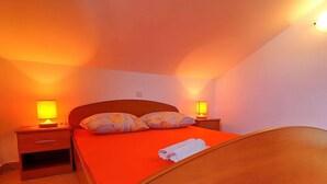 A single bed with a wooden frame and a red bedspread, flanked by two nightstands with lamps, in a room with white walls and a sloped ceiling. - Apartments Marko Peljesac - Two Bedroom Apartment With Balcony (Orebic)