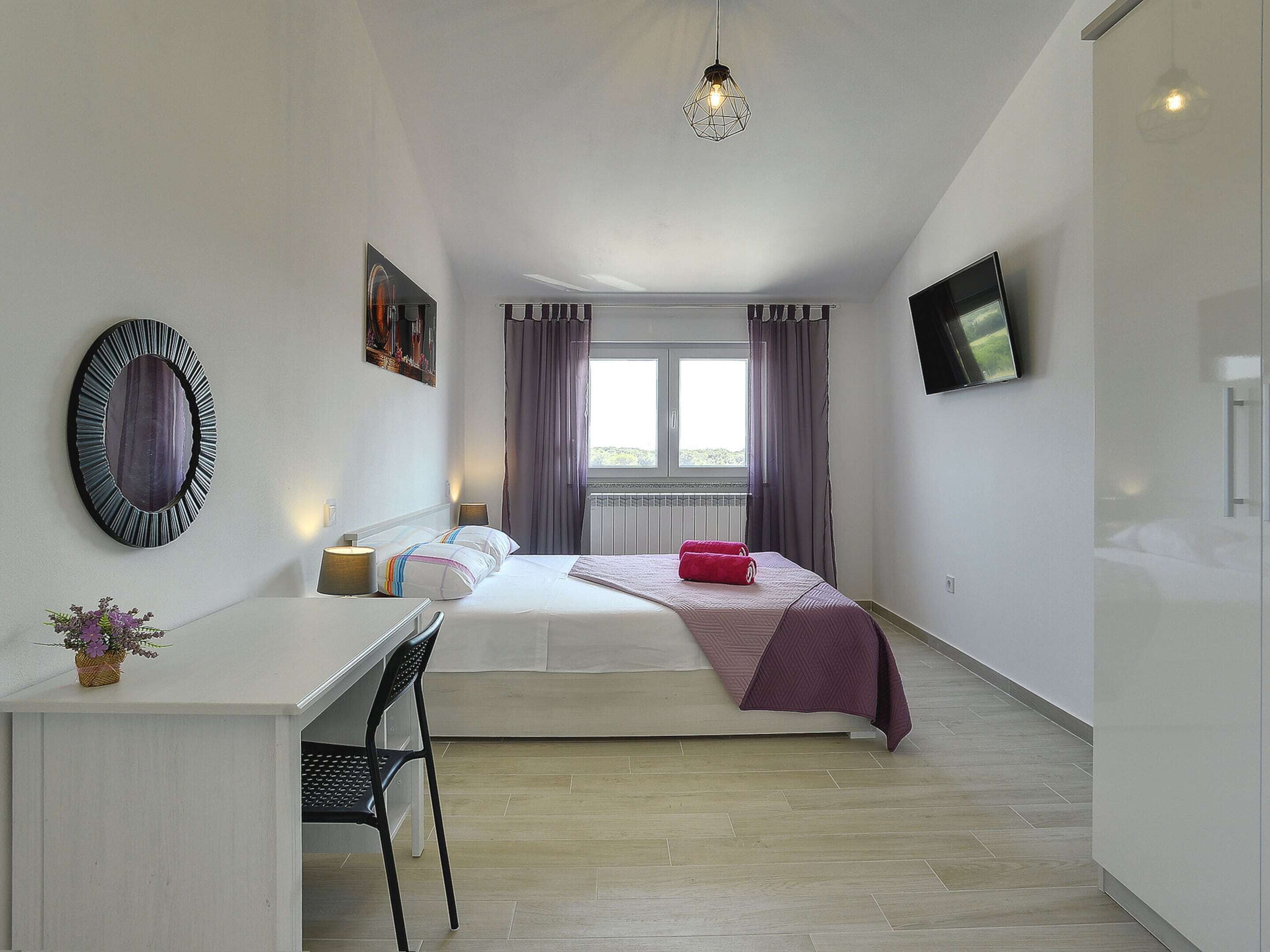 A modern hotel room with a bed, a desk, and a chair. There is a window with curtains, a wall-mounted TV, and a decorative mirror.