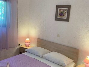 A bedroom with a bed, two pillows, a nightstand with a lamp, a framed picture on the wall, and a window with curtains. - Apartment With one Room Near City Centre (Pula)