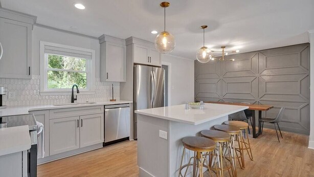 Una cocina moderna con armarios blancos, un refrigerador de acero inoxidable, una isla central con taburetes, iluminación colgante y un ventanal que aporta luz natural.