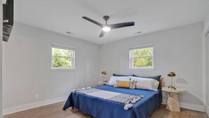 Un dormitorio con cama, dos ventanas, ventilador de techo y una mesita de noche con lámpara.