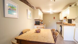 A kitchen with white cabinets, a wooden dining table, and chairs. There is a framed picture on the wall and a ceiling light fixture. - Ynys (Dyffryn Ardudwy)