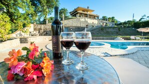 Una mesa con una botella de vino y dos copas, un arreglo floral y vista a la piscina de un edificio.