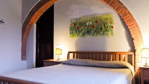 A bedroom with a wooden bed, a brick archway, a nightstand with a lamp, and a wall mural of a field with flowers. - Belvilla by OYO Vocabolo Ronda3 12 p (Parrano)