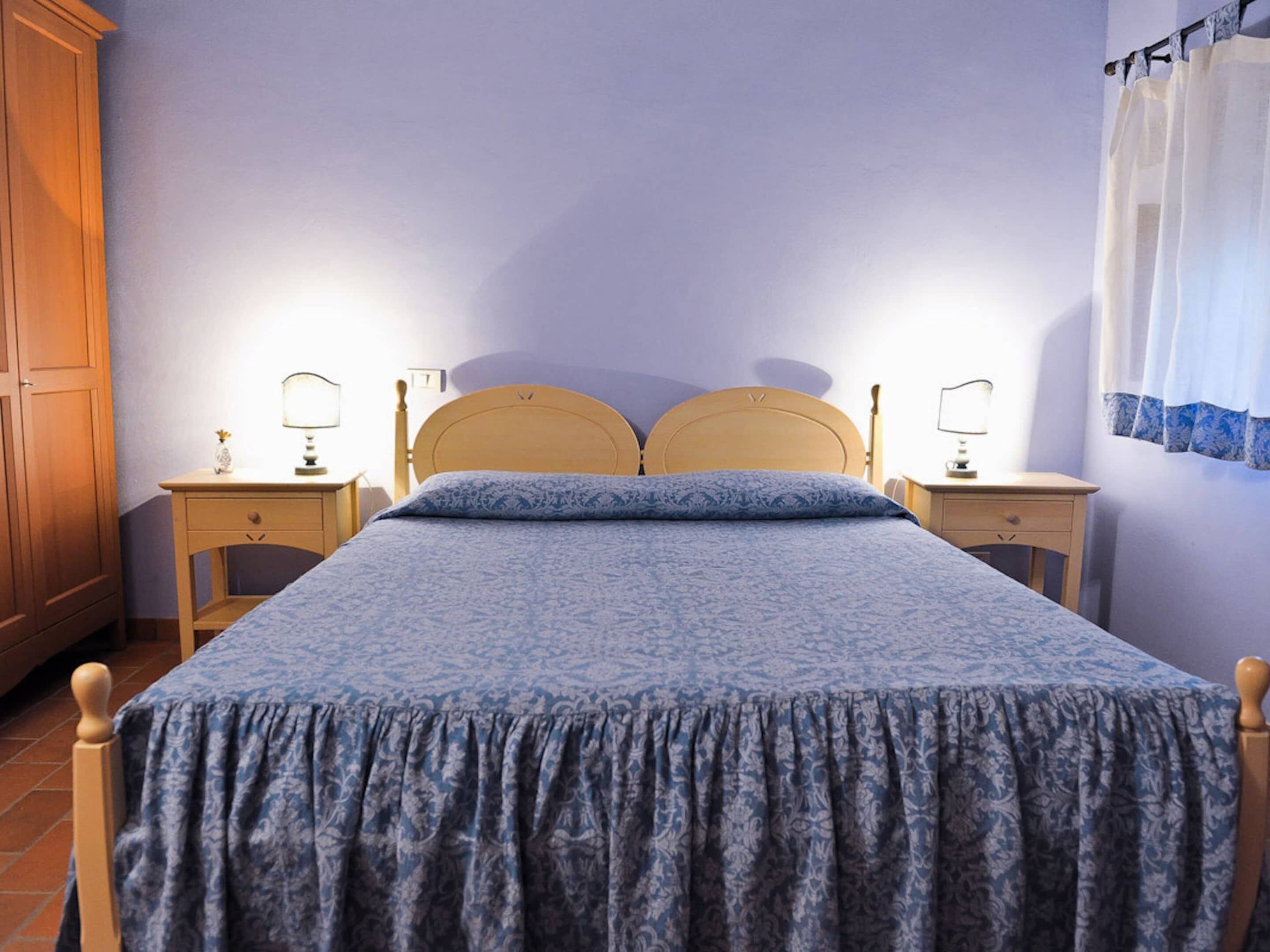 A bedroom with a bed, two bedside tables, a nightstand, a wooden wardrobe, and a window with curtains.