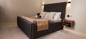 A bed with a dark headboard, a bedside table with a lamp, and a small wooden shelf with a power outlet. - Remarkable 2-bed Apartment, Dar el Salam, Tanzania (Dar es Salaam)