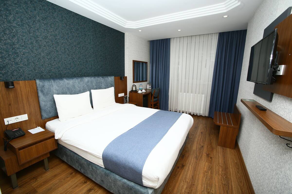 A hotel room with a large bed, a wooden nightstand, a desk, a chair, a TV mounted on the wall, and a window with curtains.