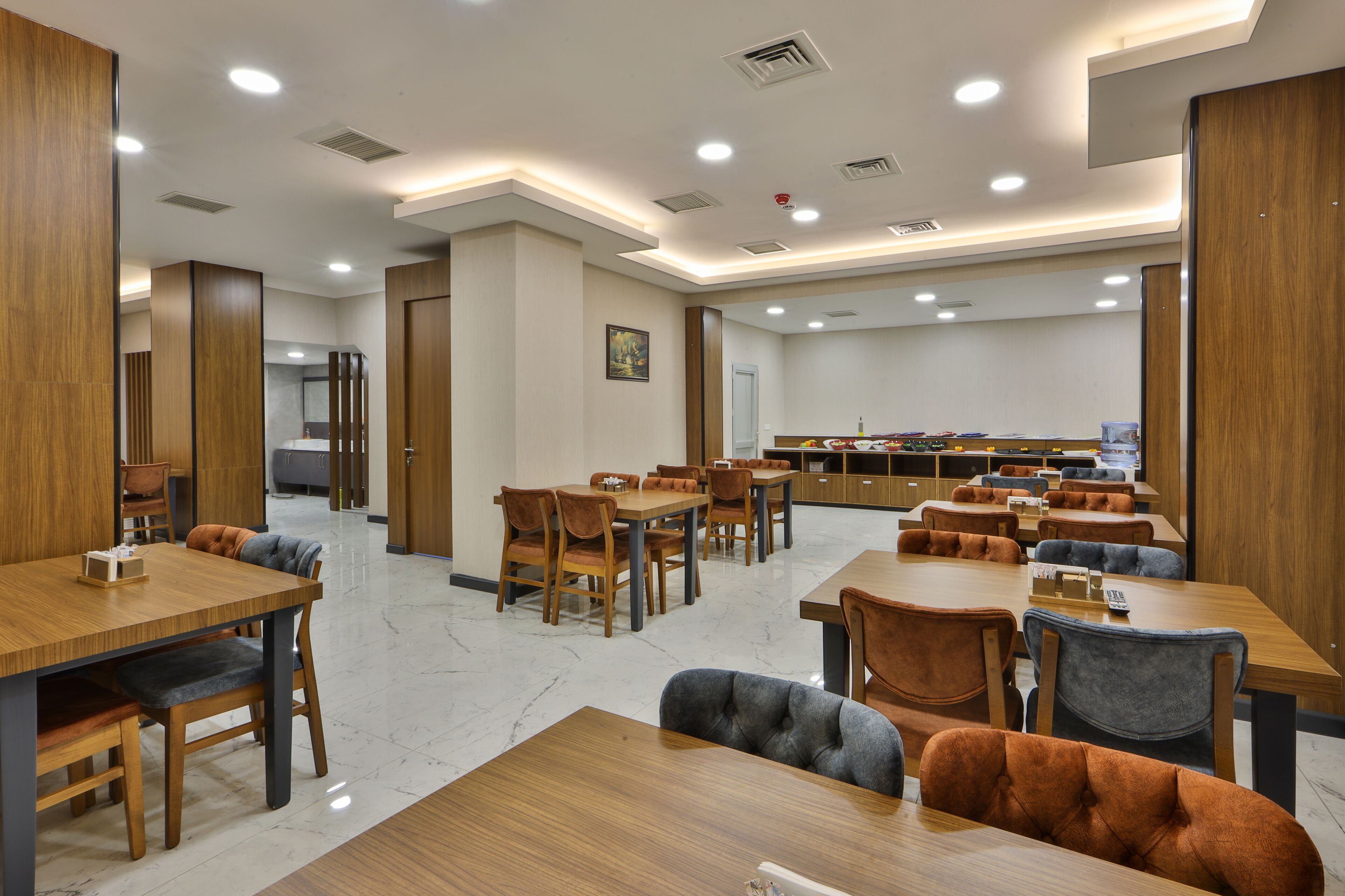 Un comedor moderno con varias mesas y sillas de madera, una cocina al fondo y un techo bien iluminado con luces empotradas.