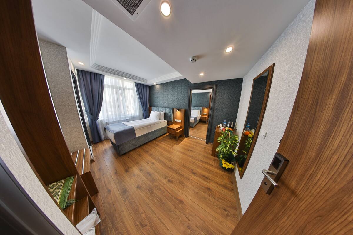 A modern hotel room with a wooden floor, a bed with a gray headboard, a bedside table, a nightstand, and a large mirror.