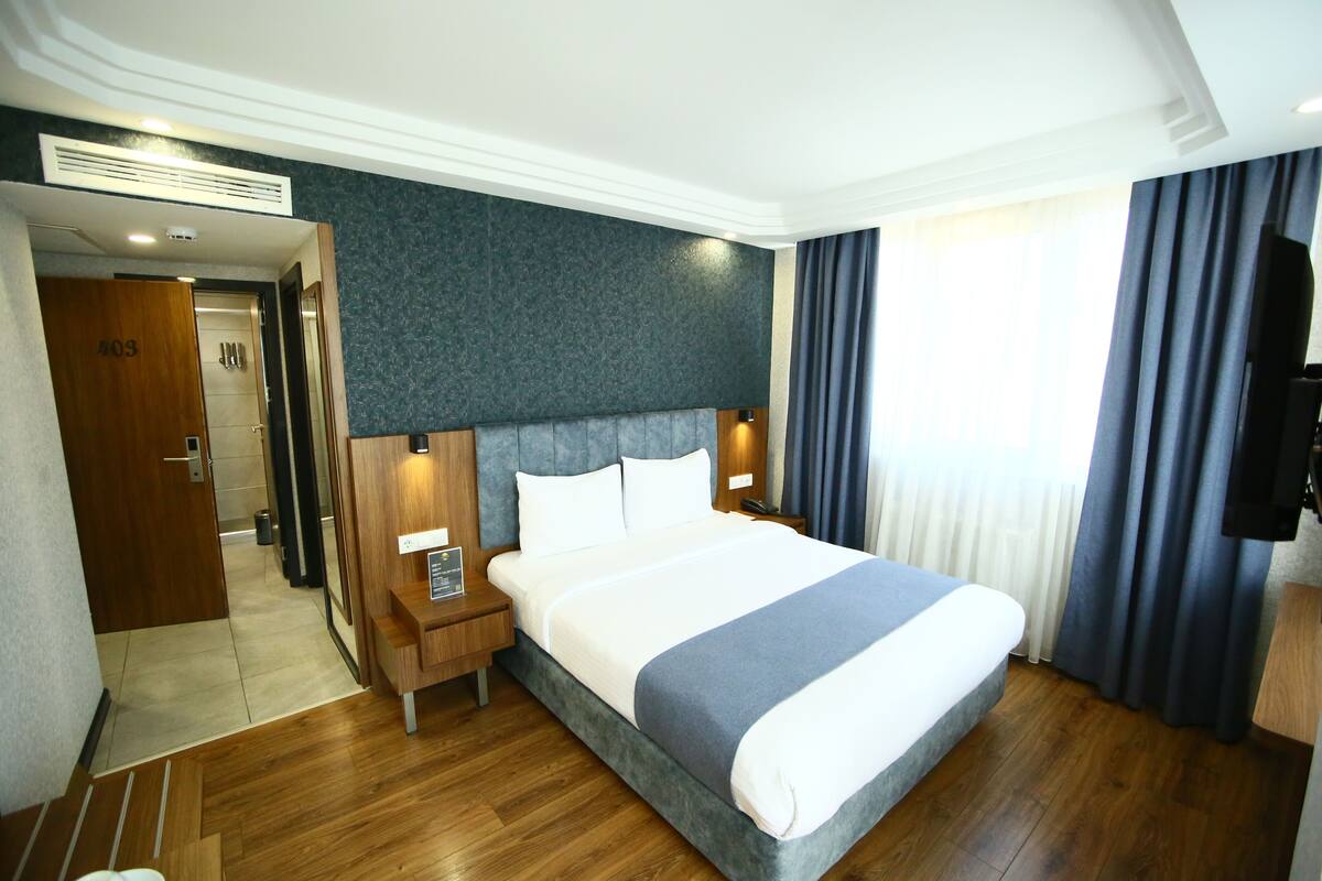 A hotel room with a large bed, wooden flooring, a dark wall, and a television on the right.