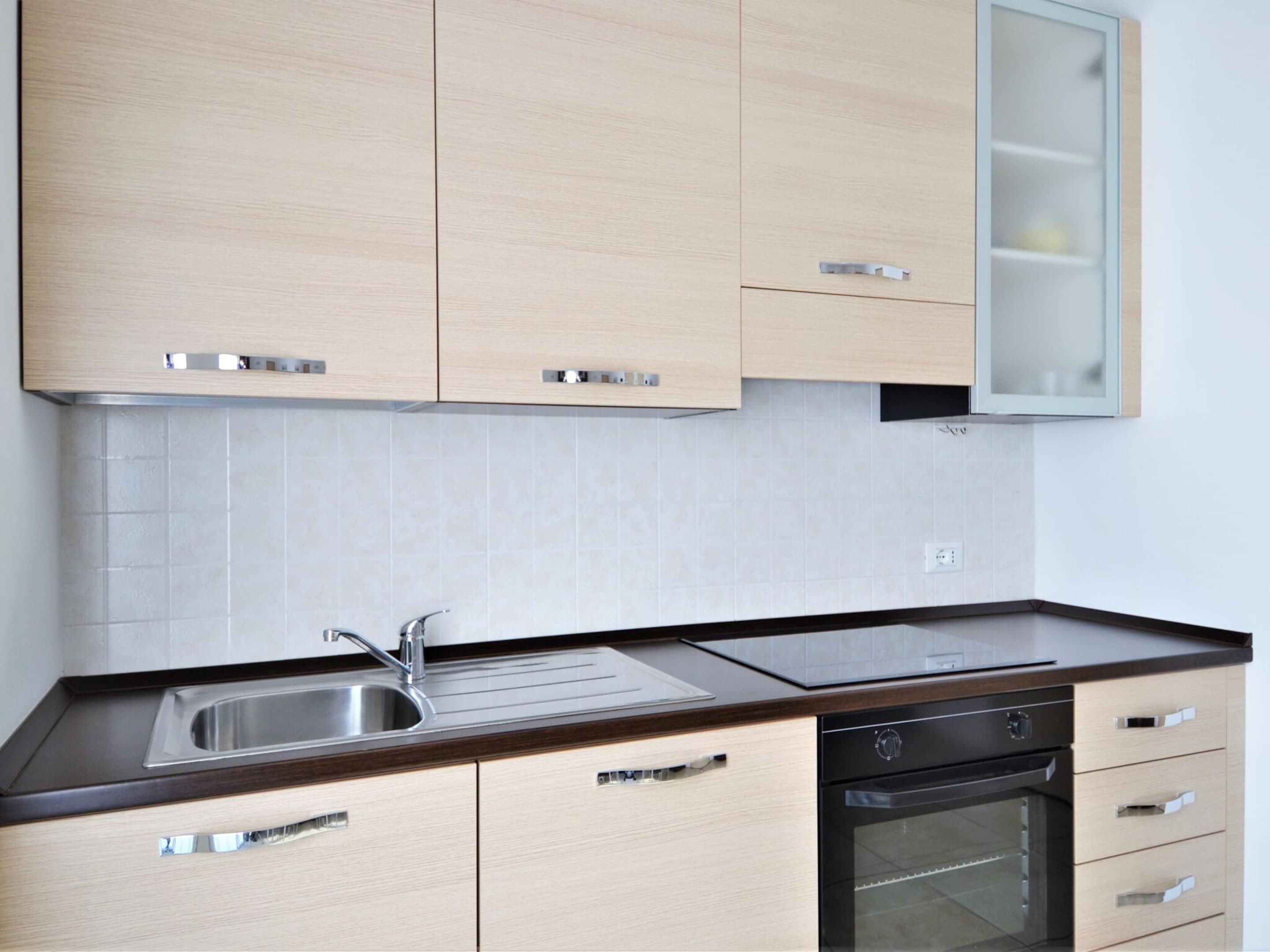A modern kitchen with light wood cabinets, a stainless steel sink, a black oven, and a glass-fronted cabinet.