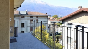 A balcony with a view of a mountain range, a building with a tiled roof, and a fenced area with greenery. - Apartment by Lake Como Near Spiaggia di Dervio (Dervio)