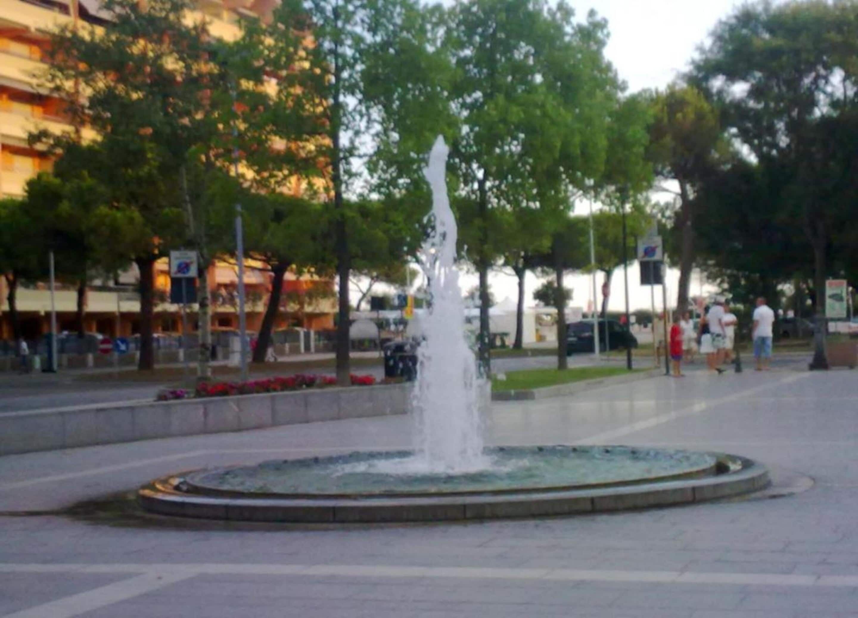 A public fountain in an urban square with trees and buildings in the background.