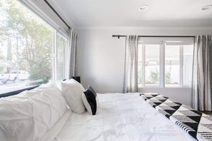 A bedroom with a large bed, white bedding, a window with curtains, and a view of the outside.
