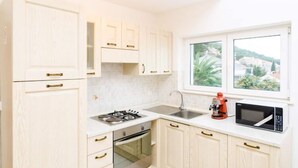 A compact kitchen with white cabinets, a gas stove, a sink, and a window with a view of greenery and buildings. - Apartments Nikola - Superior One Bedroom Apartment With Sea View (Dubrovnik)