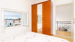 A bedroom with a large bed, a wardrobe, a window with a view of buildings, and a balcony with a railing. - Apartments Nikola - Superior One Bedroom Apartment With Sea View (Dubrovnik)
