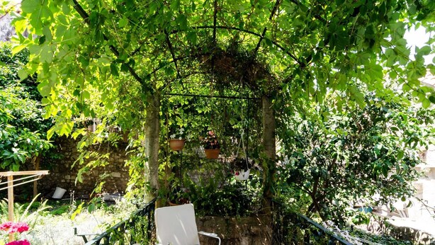 A garden patio with a white table set for dining, surrounded by lush greenery and a stone wall. - Apartment Taura - Studio Apartment With Balcony and Old City View (Dubrovnik)