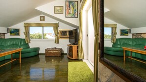 Uma sala de estar ampla com um sofá de couro verde, uma mesa de centro de madeira, uma mala antiga e uma televisão.