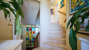 Uma escada com corrimão, uma porta que dá para um quarto com janelas de vitrais coloridos e um quadro na parede.