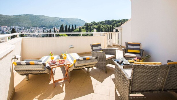 Uma varanda com móveis de vime, uma mesa com frutas e bebidas, e vista para uma cidade e colinas ao fundo.