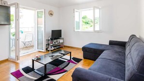 Ein modernes Wohnzimmer mit grauem Sofa, einem Glastisch und einem Fernseher auf einem Schrank. Zwei Fenster sorgen für natürliches Licht und bieten Blick ins Grüne.