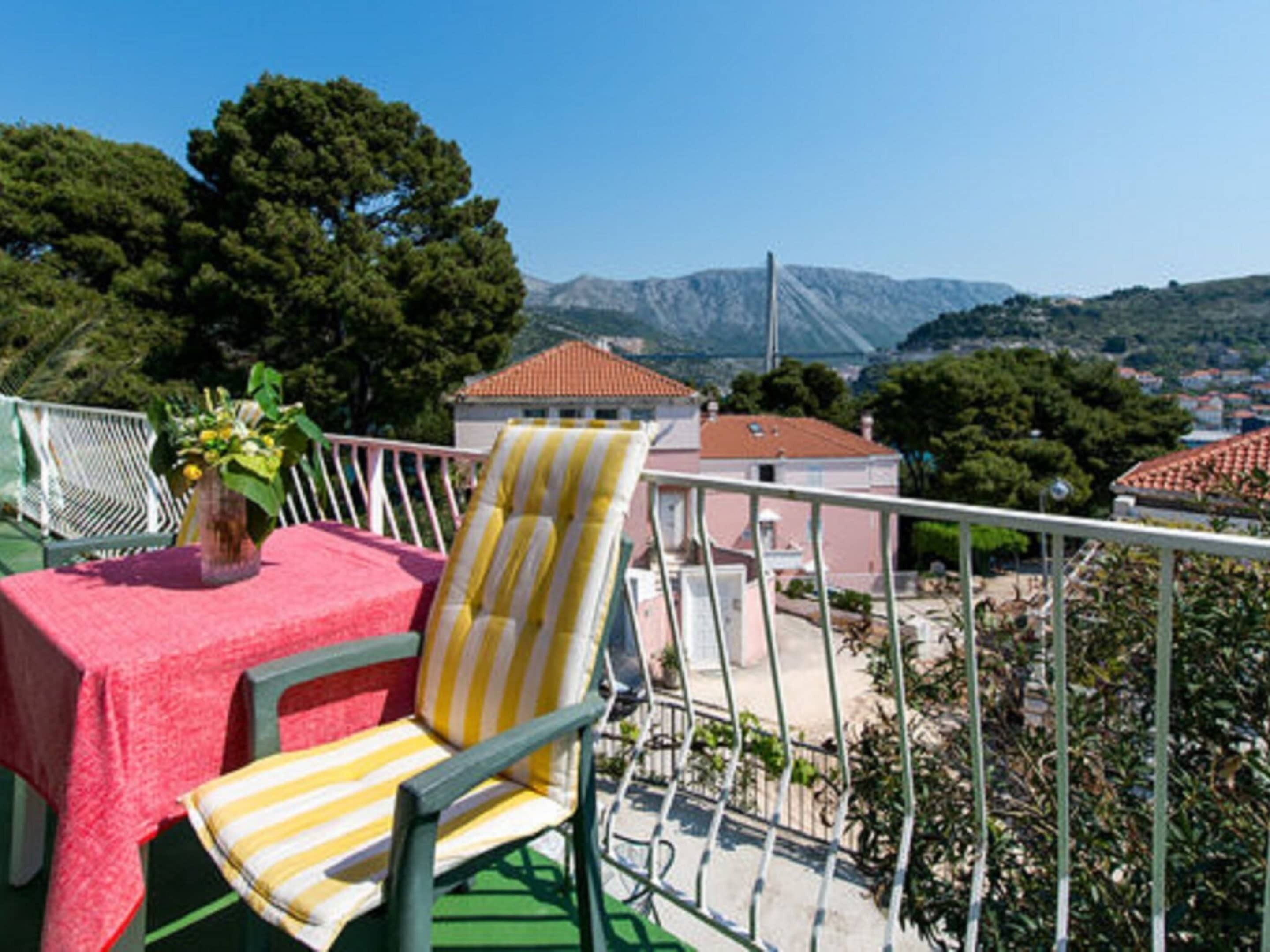 A balcony with a table covered in a red tablecloth and a striped chair, overlooking a town with buildings and mountains in the background.