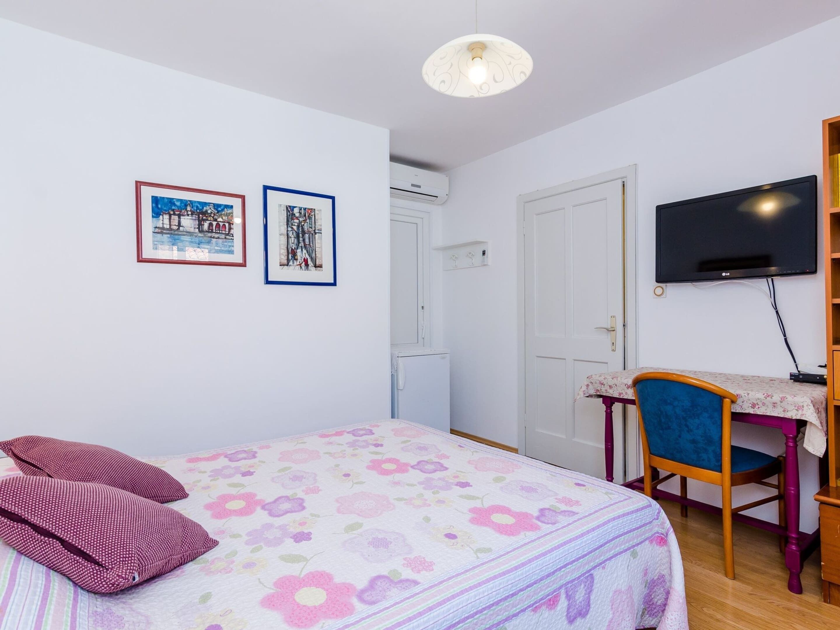 Een slaapkamer met een bed, een bureau, een stoel, een televisie en twee afbeeldingen in een lijst aan de muur.