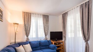 Ein Wohnzimmer mit einem blauen Sofa, einem Fernseher auf einem Holzgestell und einem kleinen Tisch mit einer Obstschale.