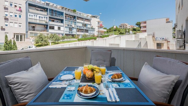 Ein Balkon mit Frühstücksangebot, darunter Croissants, Obst und Orangensaft. Im Hintergrund sind moderne Gebäude und ein klarer Himmel zu sehen.