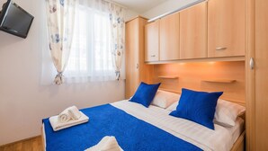 Ein Hotelzimmer mit Bett, Bettwäsche in Blau und Weiß, einem Holzschrank, einem an der Wand montierten Fernseher und einem Fenster mit transparenten Vorhängen.