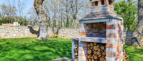 Un barbecue en pierre doté d’une cheminée, entouré d’un muret en pierre et d’une zone herbeuse avec des arbres.