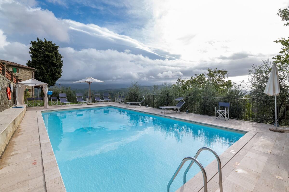 Una piscina con agua cristalina azul, rodeada de sillas de descanso y sombrillas, con vistas a montañas al fondo.