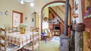 Un coin repas chaleureux avec une nappe à carreaux, des chaises en bois et un vase rempli de fleurs. Un escalier mène à un salon équipé d’une télévision et d’une cheminée.