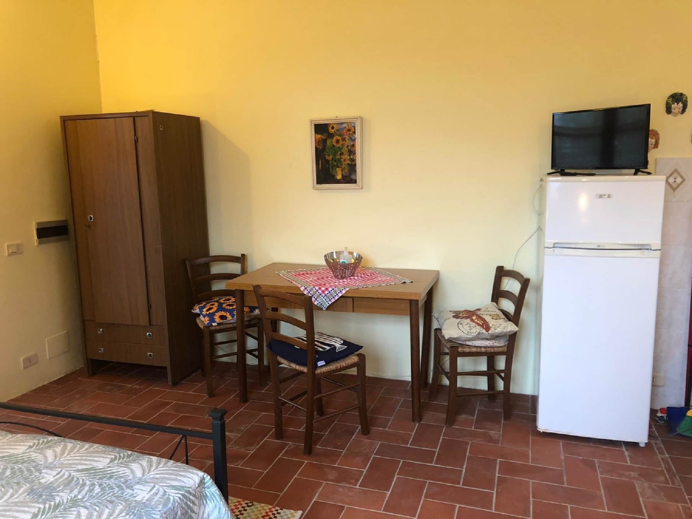A small room with a wooden table, two chairs, a white refrigerator, a wooden wardrobe, and a television on top of the fridge.