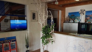 Aula, jossa on puinen vastaanottotiski, televisio, jolta näkyy kaunis maisema, huonekasvi ja seinä, jossa on meriaiheinen maalaus.