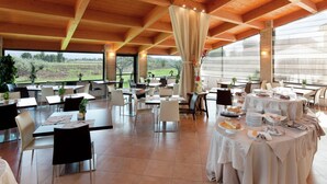Un comedor amplio con techos altos y ventanas grandes, que ofrece vistas al campo. Las mesas están dispuestas con manteles blancos y diferentes platos.