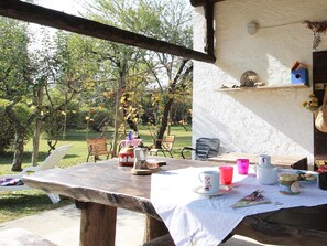 An outdoor dining area with a table set for two, surrounded by chairs, and a view of a garden with trees and grass.
