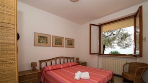 Een slaapkamer met een bed, een stoel, een nachtkastje en een raam met uitzicht op bomen.