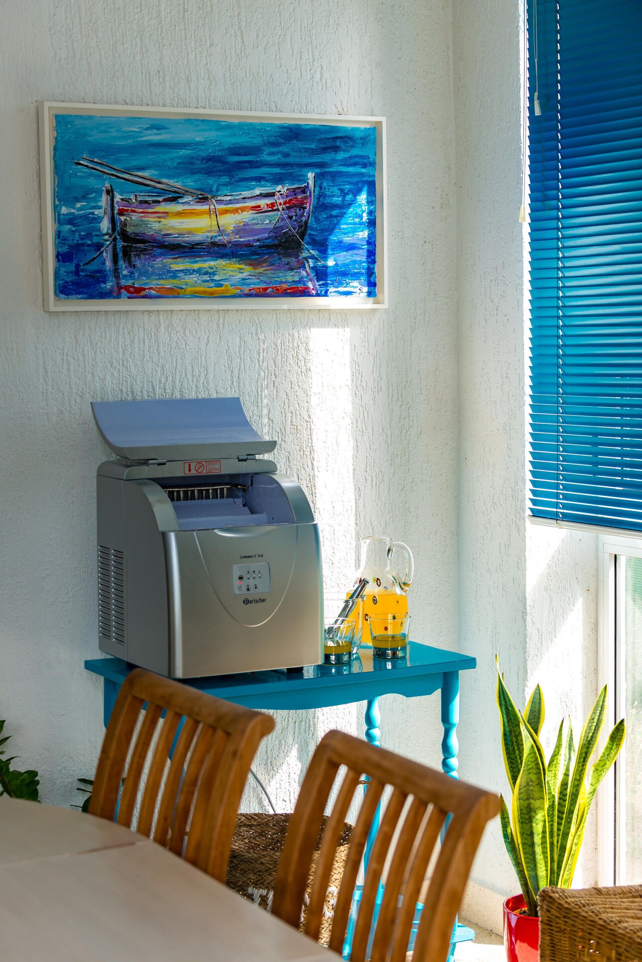 Um quarto com uma mesa, cadeiras, uma impressora, um quadro e uma janela com persianas.
