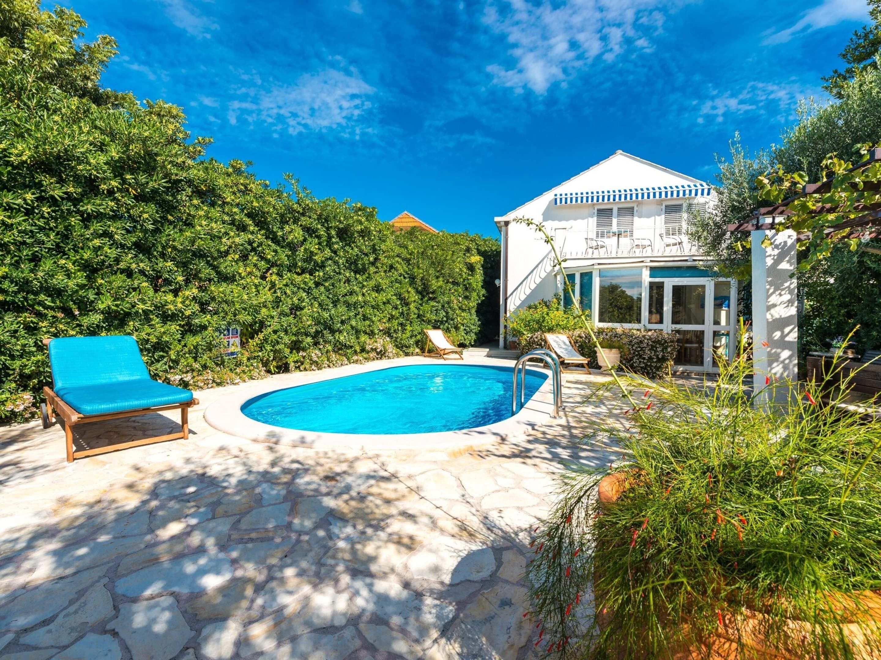 Piscina com espreguiçadeiras, um pátio de pedra e um edifício branco com telhado azul.