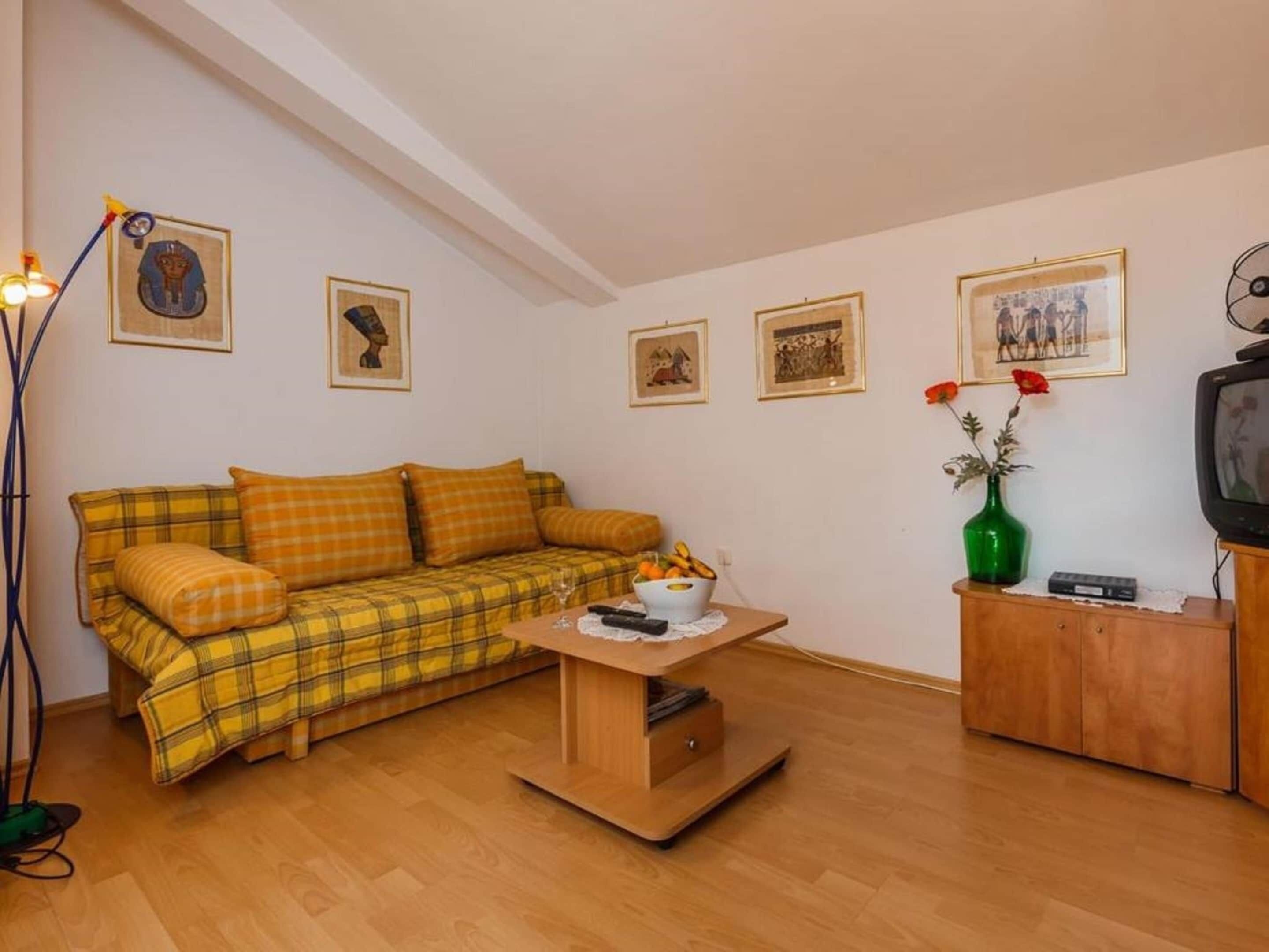 A cozy living room with a plaid sofa, a small wooden table, a green vase with flowers, and a television on a wooden cabinet.