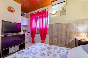 Een slaapkamer met een bed, een televisie, een kledingkast, een raam met gordijnen, een airconditioner en een aan de muur hangend schilderij.